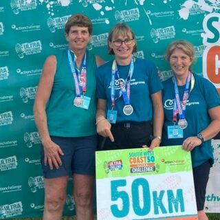 3 Ladies wearing Love Musgrove-branded, light teal-cooloured t-shirts and wearing medals. Holding a 50km sign, whilst a large banner saying 'Finisher. South West Coast 50 Ultra Challenge' hangs behind them.