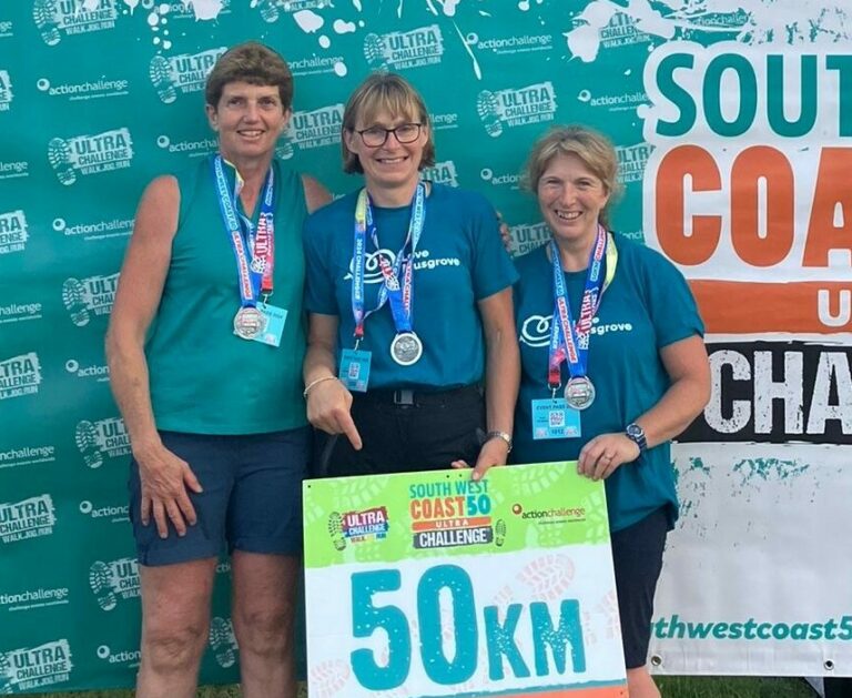 3 Ladies wearing Love Musgrove-branded, light teal-cooloured t-shirts and wearing medals. Holding a 50km sign, whilst a large banner saying 'Finisher. South West Coast 50 Ultra Challenge' hangs behind them.