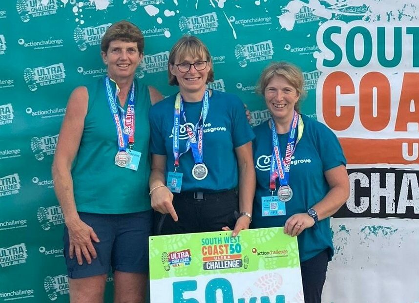 3 Ladies wearing Love Musgrove-branded, light teal-cooloured t-shirts and wearing medals. Holding a 50km sign, whilst a large banner saying 'Finisher. South West Coast 50 Ultra Challenge' hangs behind them.