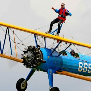 A blue and yellow plane flying in in a grey and cloudy sky, and a man is strapped and standing on the upper wings of the plane