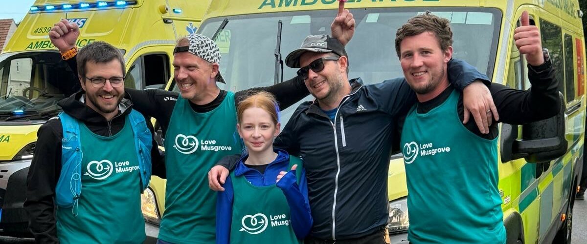 Four men and a young girl wearing Love Musgrove vests celebrate finishing an ultramarathon in front of lit up ambulances